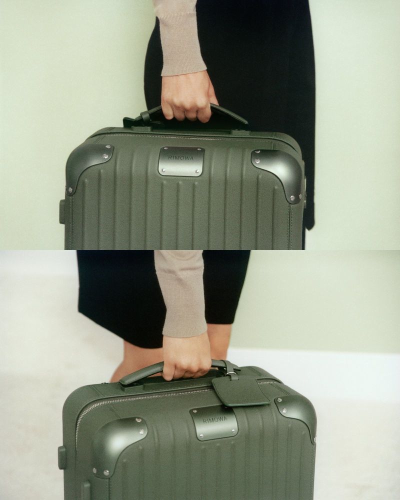 Close-up of a person carrying a dark green Rimowa suitcase with a brown handle and label in an indoor setting with a light green background.