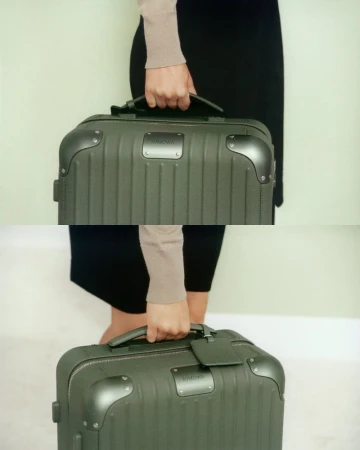 Close-up of a person carrying a dark green Rimowa suitcase with a brown handle and label in an indoor setting with a light green background.