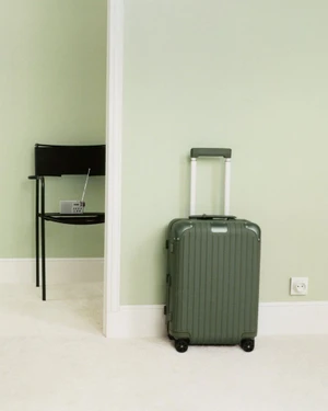 Green wheeled suitcase standing against a pale green wall next to a white electrical outlet in an interior setting