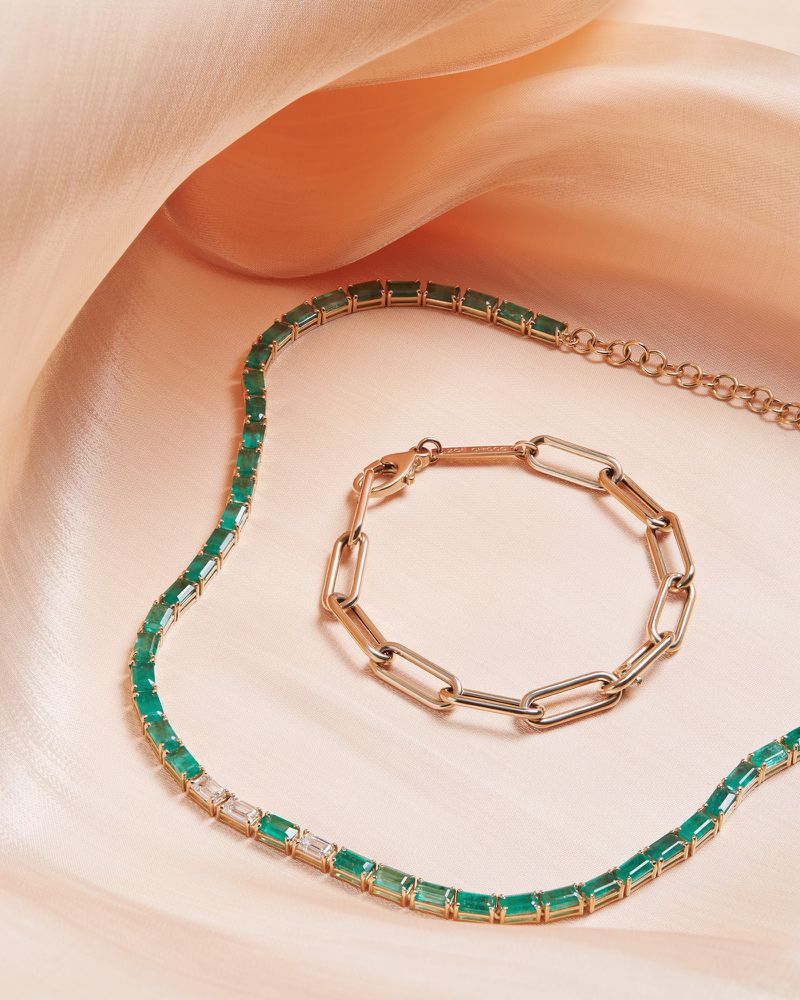 Emerald and gold necklace and gold link bracelet on a soft peach-colored satin fabric backdrop in a studio setting. Jewelry photography.