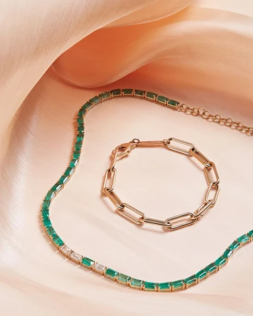 Emerald and gold necklace and gold link bracelet on a soft peach-colored satin fabric backdrop in a studio setting. Jewelry photography.
