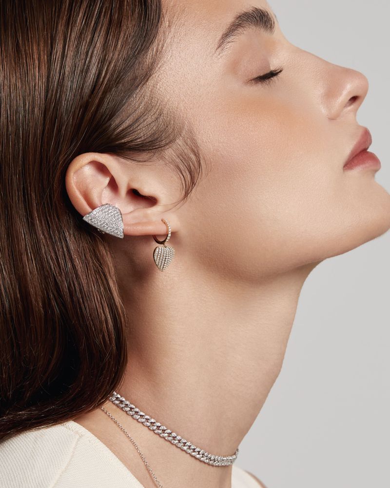 Woman showcases modern jewelry: geometric ear cuff, heart earring, chain necklaces against a neutral backdrop, emphasizing style and elegance.
