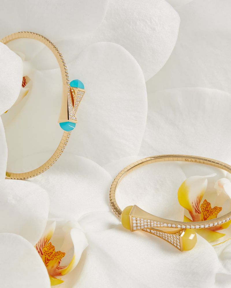 Close-up of two gold bracelets with light blue and yellow stone accents, set against a soft backdrop of white orchid petals.