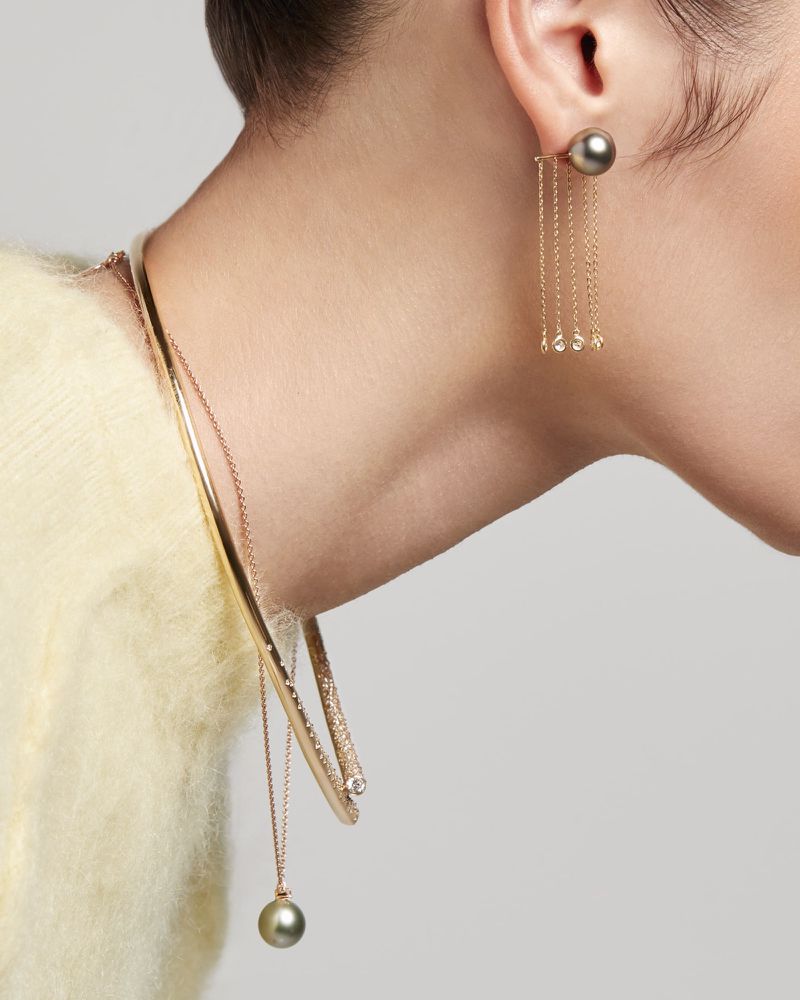 Close-up studio shot of an adult female wearing a yellow sweater, gold choker necklace and earrings with pearl drops against a gray background.