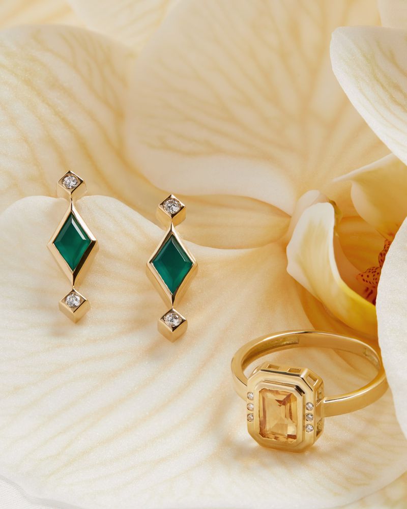 Elegant gold jewelry set featuring emerald earrings and a yellow gemstone ring, displayed against a soft, textured cream background.