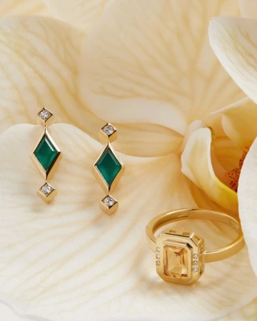 Elegant gold jewelry set featuring emerald earrings and a yellow gemstone ring, displayed against a soft, textured cream background.