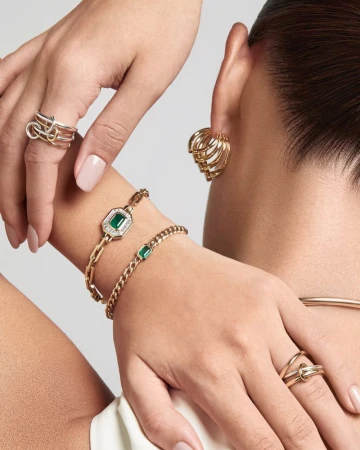 Close-up of a woman showcasing fine jewelry including gold rings, emerald bracelets, and sculptural earrings for Harvey Nichols