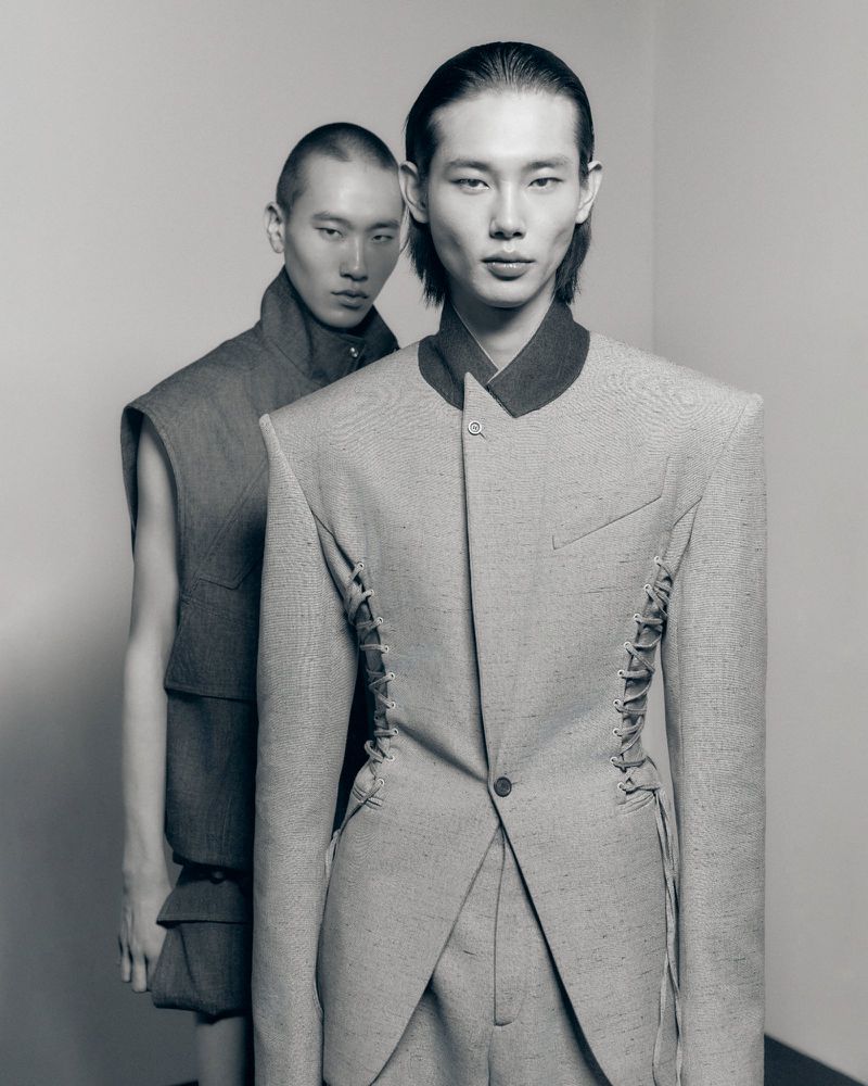 Two Asian male models in avant-garde fashion, one in a tailored suit with unique lacing, the other in a layered sleeveless vest.