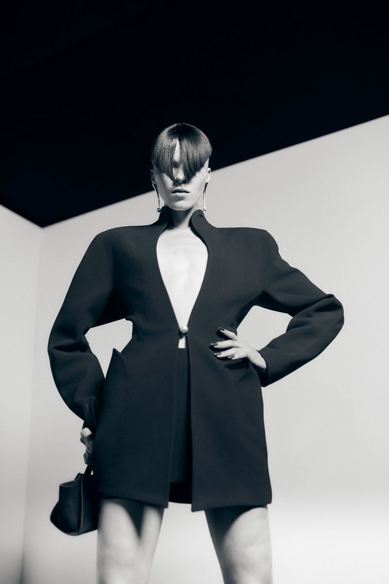 Monochrome fashion shot of adult female posing indoors wearing an oversized black blazer, dark skirt, earrings, and holding a black purse.