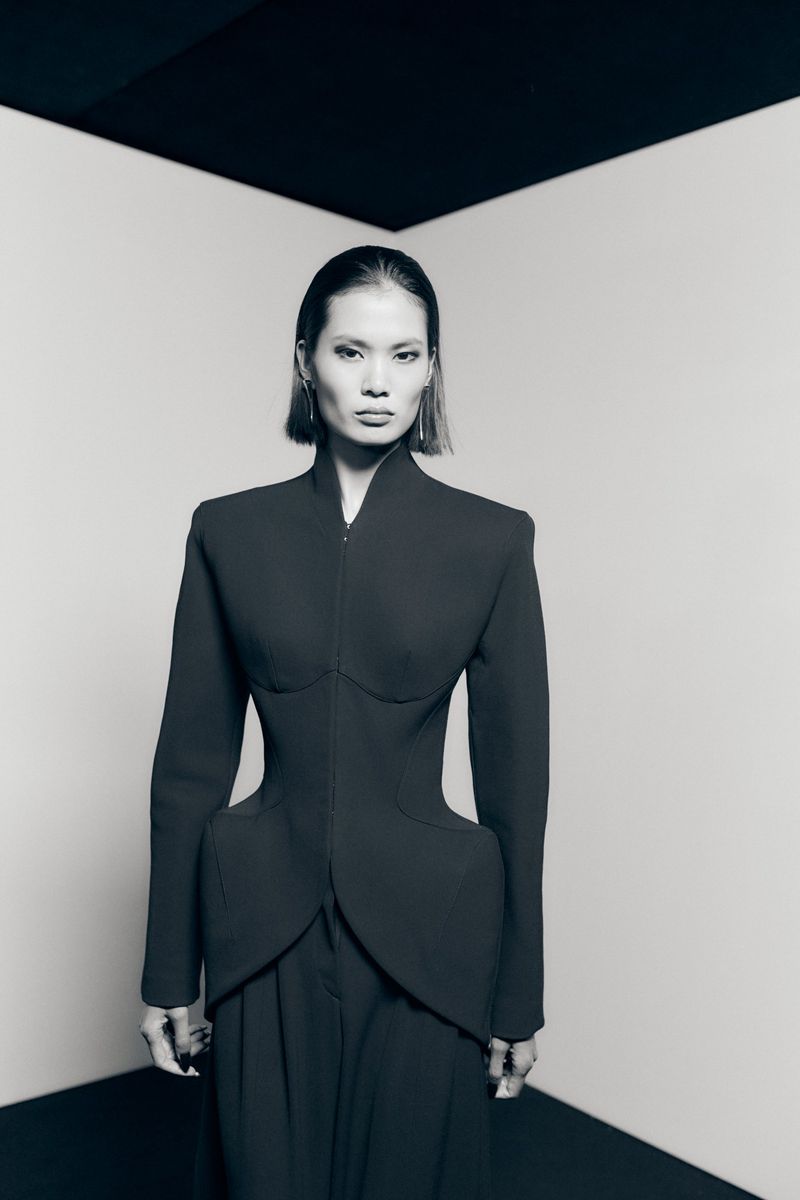 Model in black structured tailored jacket with architectural padded hips against a neutral corner backdrop.