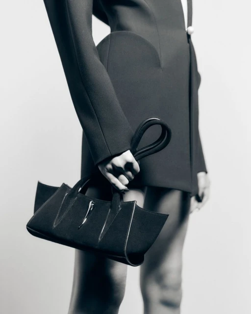 Fashion shot of a black handbag with chrome hardware, held by a person wearing a black blazer, presented against a neutral background.