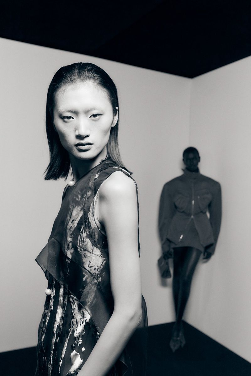 Two women in a studio setting. The woman in the foreground wears a sleeveless top. The background woman wears a jacket and leggings.