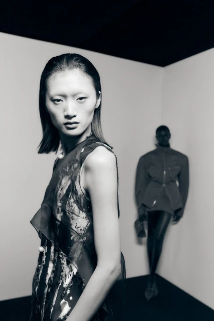 Two women in a studio setting. The woman in the foreground wears a sleeveless top. The background woman wears a jacket and leggings.