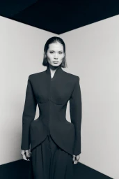 Model in black structured tailored jacket with architectural padded hips against a neutral corner backdrop.