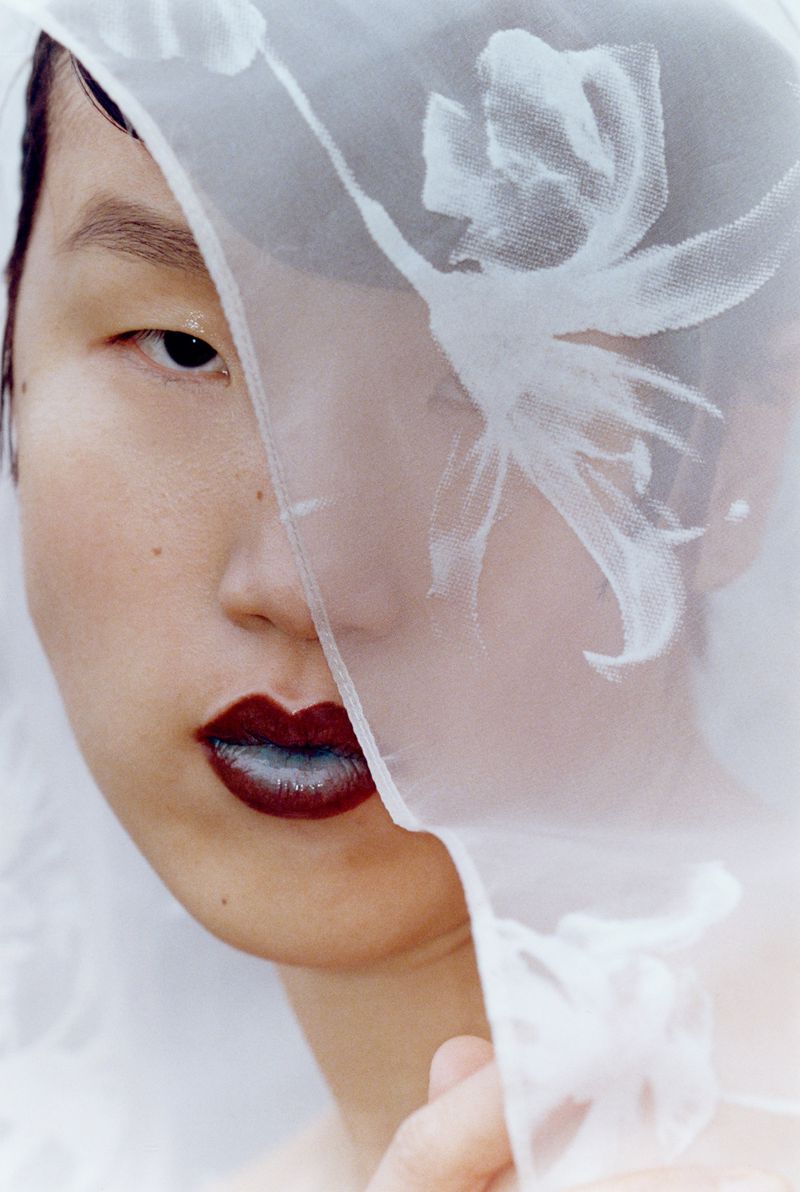 Close-up of a stylish adult with dark lipstick, partially covered by a sheer white fabric with floral print, set against a light background.