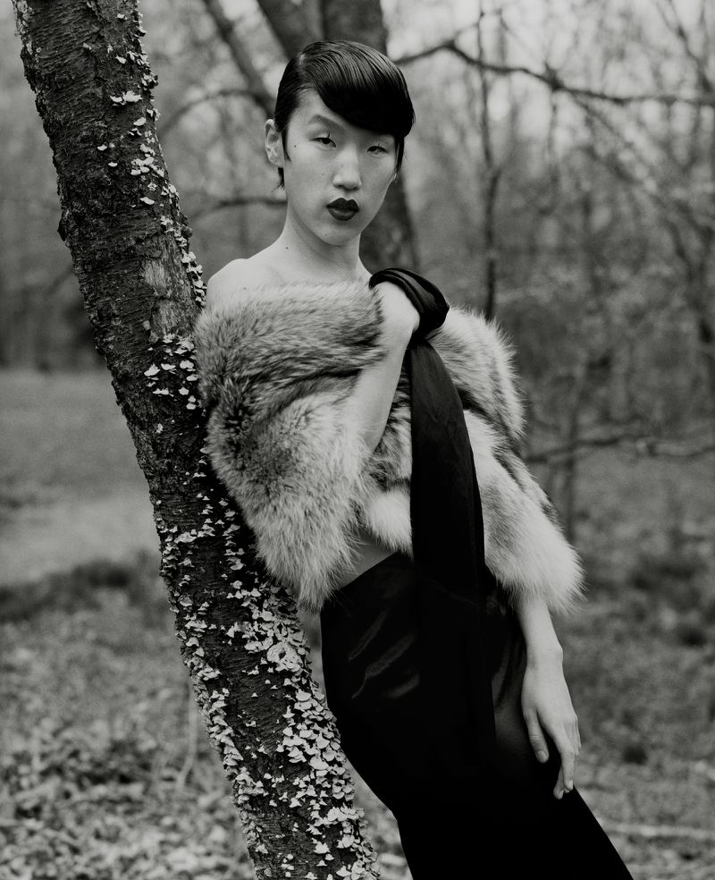Portrait of a woman in a fur stole leaning against a tree in a forest setting. Black and white photography with shallow depth of field.