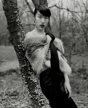 Portrait of a woman in a fur stole leaning against a tree in a forest setting. Black and white photography with shallow depth of field.