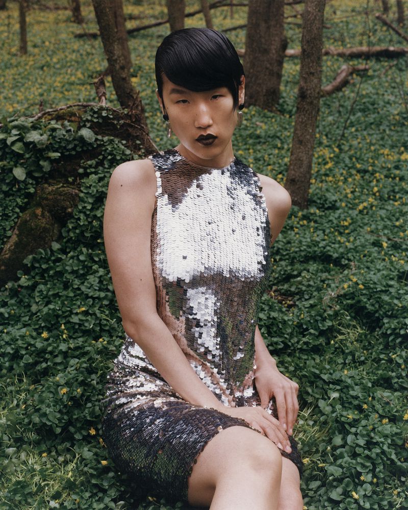 Elegant woman with sleek black hair and dark lipstick poses in a shimmering sleeveless dress in a forest setting with dappled sunlight.