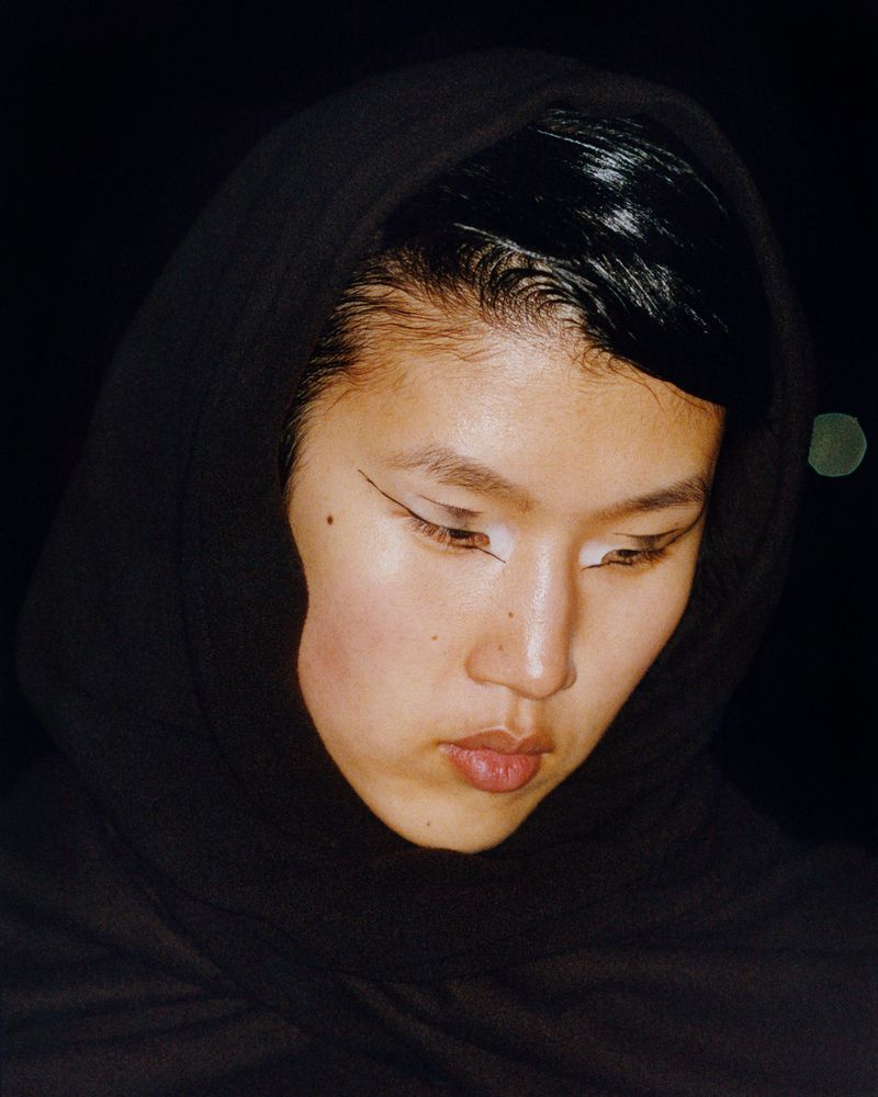Close-up portrait of an Asian woman with black winged eyeliner and a dark hood, looking down against a dark background.