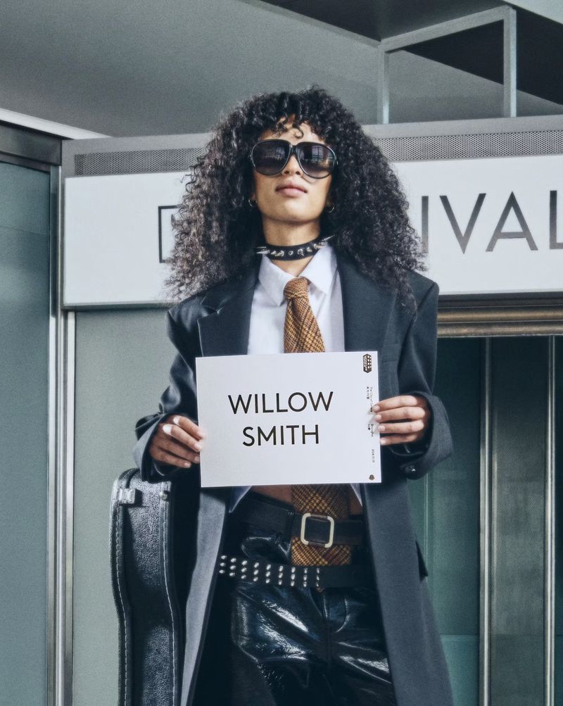 Adult woman in a black coat holds a sign that reads "Willow Smith" in an indoor location with a suitcase next to her.