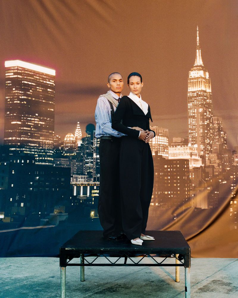A fashion shot captures a man and woman posing on a small platform with a large backdrop of a nighttime city skyline featuring the Empire State Building.