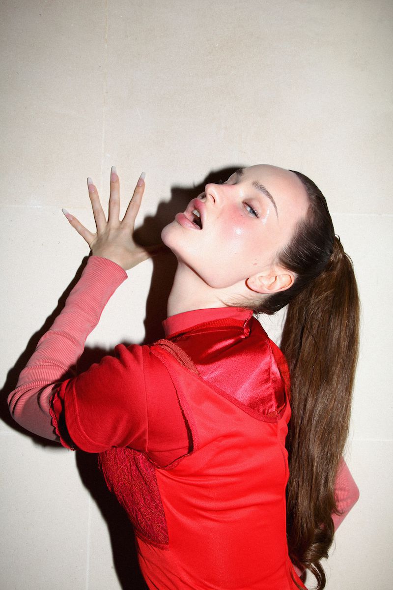 Woman in red turtleneck and long ponytail looks up with open mouth, long nails, and hand raised against a plain light-colored wall.