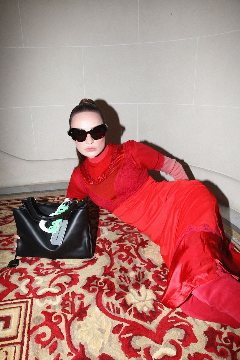 A woman in a red dress and sunglasses lounges on a patterned rug with a black designer bag, creating a fashionable indoor portrait.