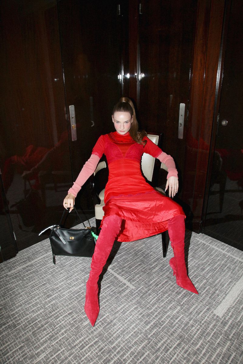 Fashionable woman in a red dress and matching thigh-high boots poses in a modern interior with dark wood panels and a patterned floor.