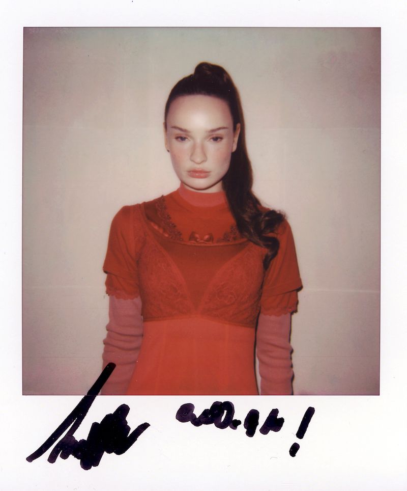 Portrait of a young woman in a red dress with lace detailing, standing indoors with a ponytail hairstyle against a plain background.