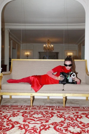 Kim Petras posing on a beige sofa wearing a red dress, sunglasses, and holding a black handbag for Balenciaga.