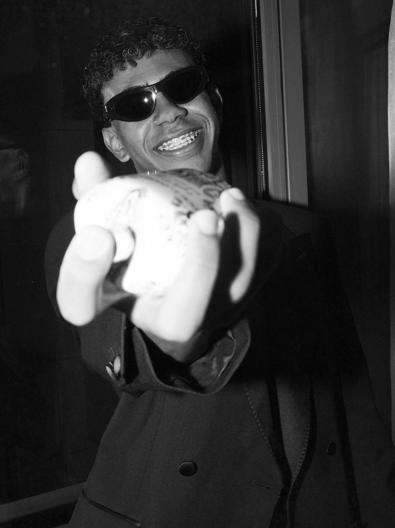 Portrait of a young adult male wearing sunglasses and a suit jacket holding an object, photographed indoors in black and white.