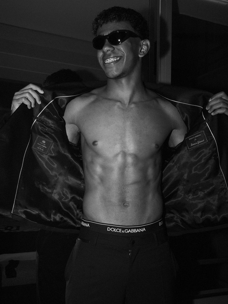 Young man in sunglasses and underwear shows off his abs while holding open his blazer in a dimly lit indoor environment.