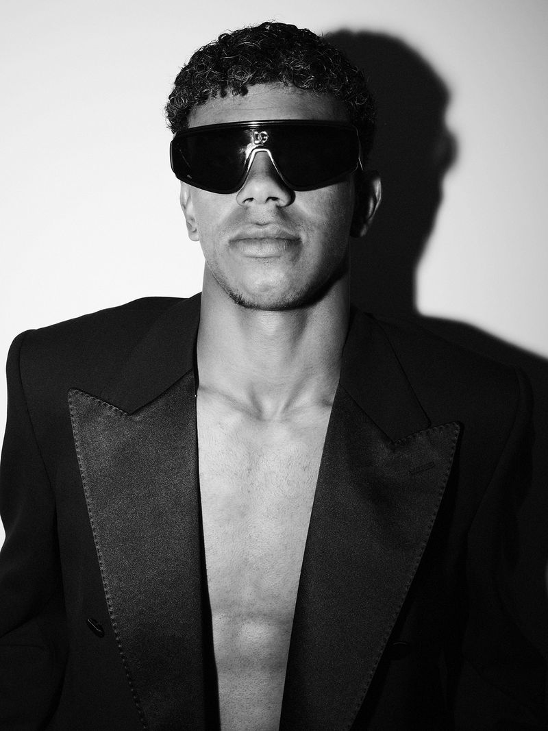 Portrait of Lamine Yamal wearing dark sunglasses and a black blazer against a light wall with a sharp shadow