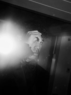 Monochrome portrait of young man in black tuxedo with bowtie, shielding eyes from a bright light flare.