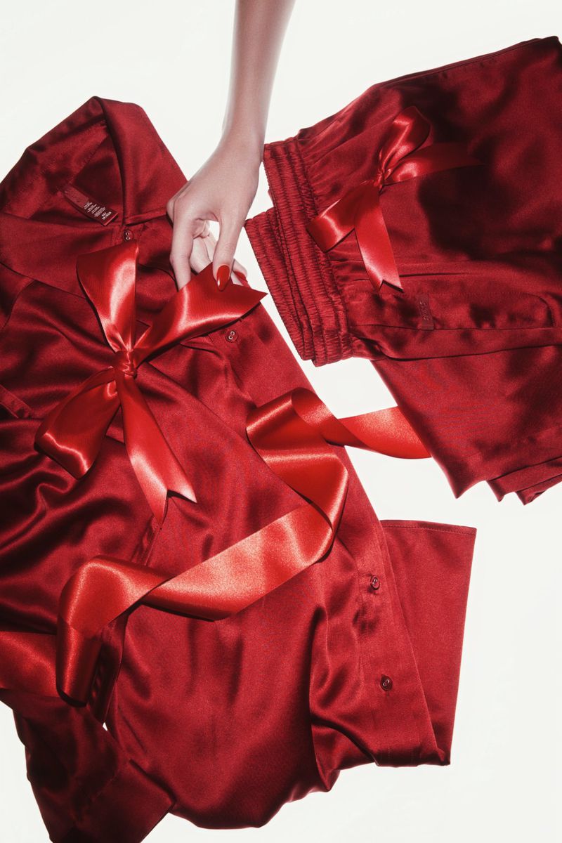 A hand holds red satin pajamas tied with a ribbon, showcasing luxurious loungewear in a studio setting against a plain white background.