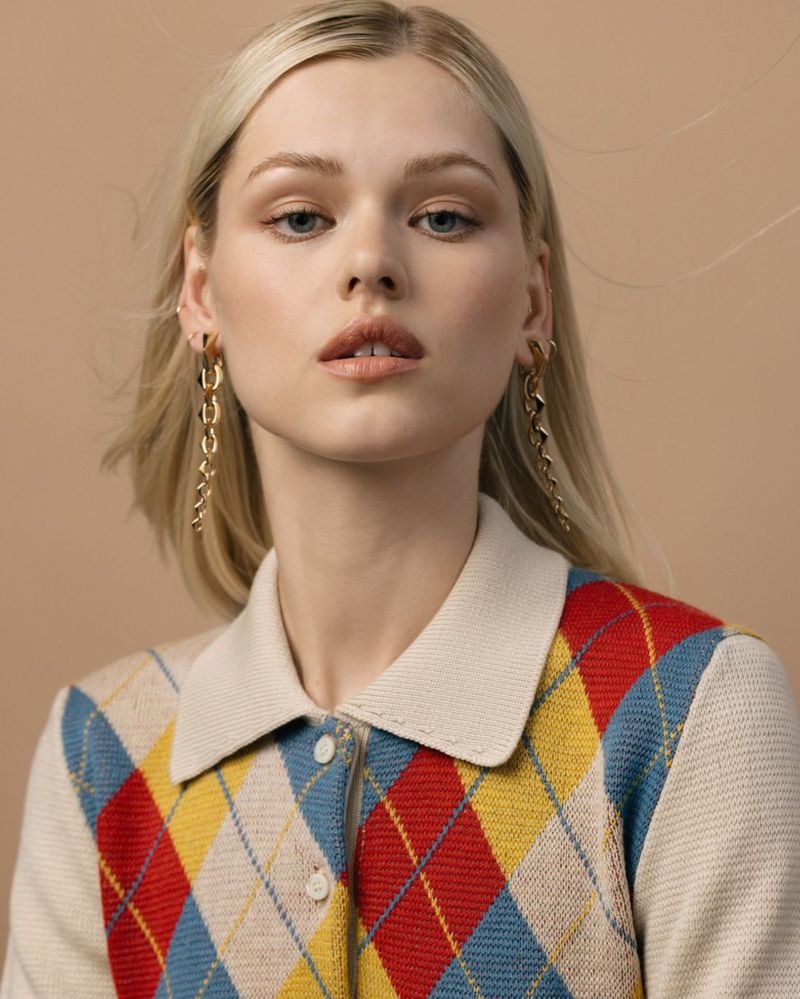Close-up shot of a young adult woman with blonde hair, wearing a red, blue, and yellow argyle sweater and gold chain earrings.