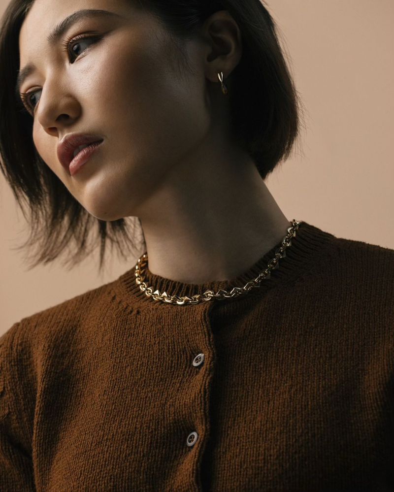 Close-up of an Asian woman with short dark hair and neutral expression. She wears a brown cardigan and gold chain necklace.