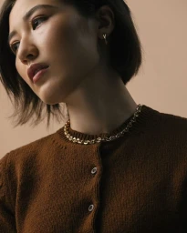 Close-up portrait of a woman wearing a brown knitted cardigan and gold chain necklace against a neutral backdrop