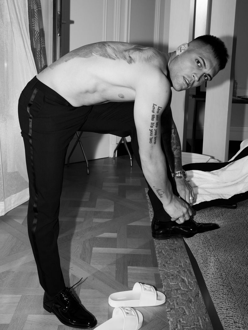 A shirtless, fair-skinned male tying his patent leather dress shoe indoors on a rug near sandals, wearing black pants and jewelry.