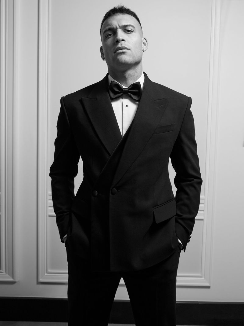 Black and white portrait of an adult male in a black tuxedo with a bow tie standing indoors in front of a textured wall.