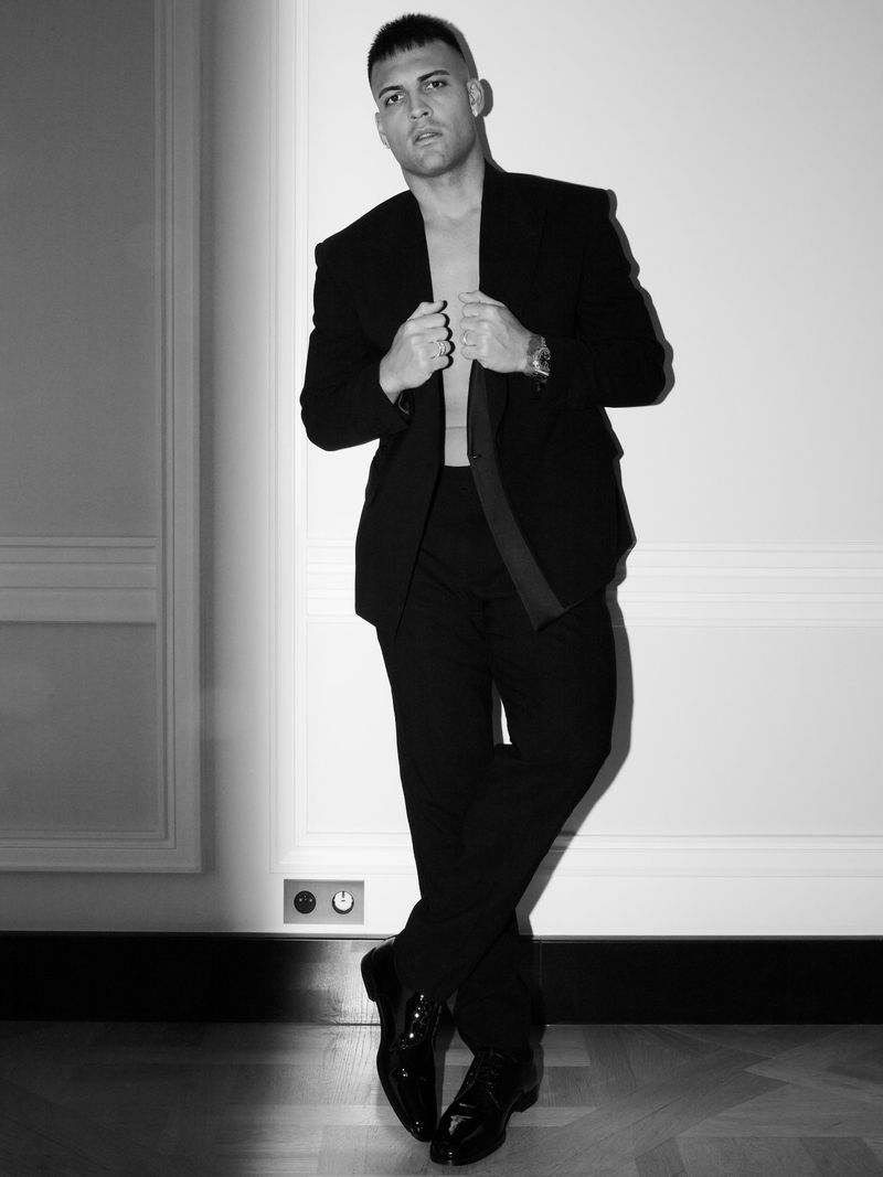 Monochrome studio portrait of a stylish adult male in an open black suit jacket and trousers posing against a white wall.