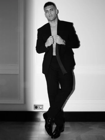 Monochrome studio portrait of a stylish adult male in an open black suit jacket and trousers posing against a white wall.