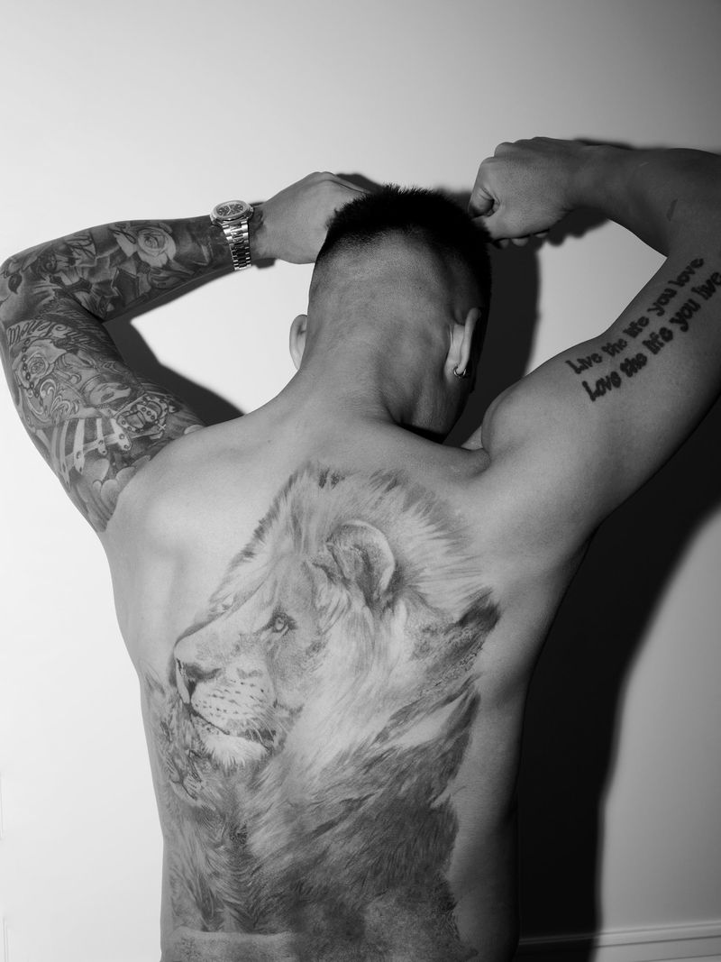 Monochrome portrait of a shirtless adult male with a lion tattoo on his back, arms raised behind his head, watch on the wrist.