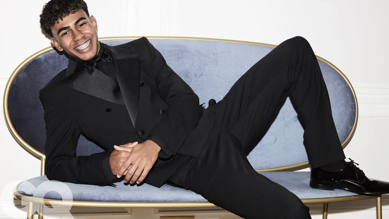 Portrait of a young man with braces in a black tuxedo lying on a light blue velvet chaise lounge in front of a white wall.