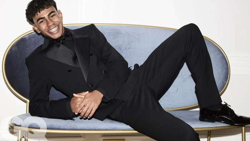 Portrait of a young man with braces in a black tuxedo lying on a light blue velvet chaise lounge in front of a white wall.