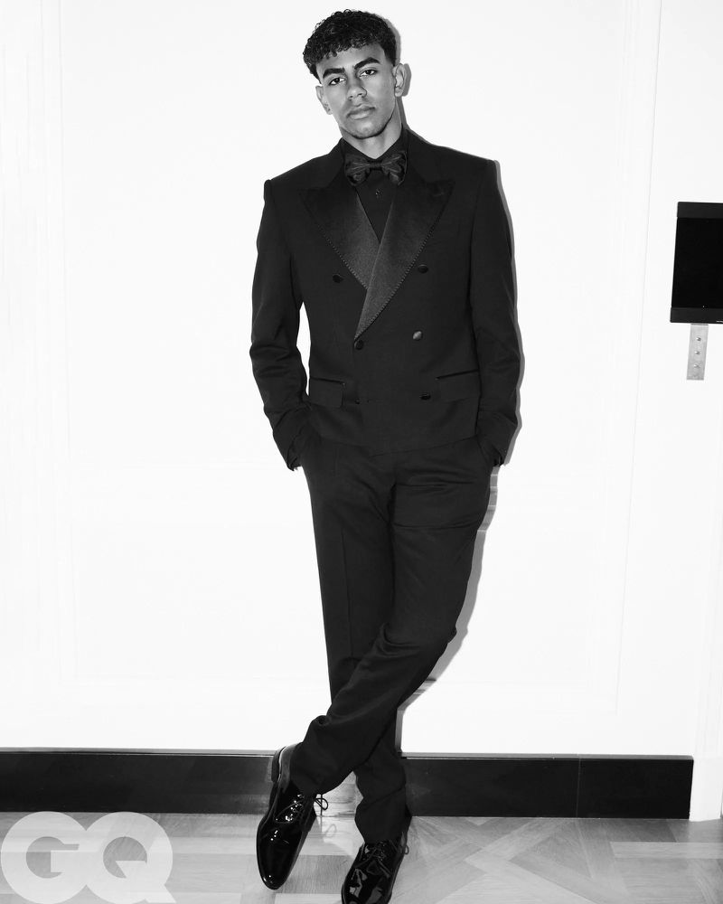 Portrait of an adult male in a black double-breasted tuxedo with a bow tie, posing against a bright white wall with dark accent molding.