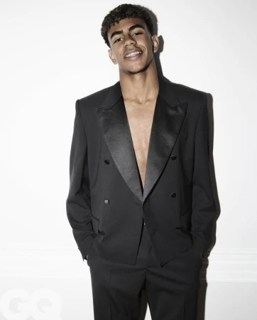 Portrait of a young adult male wearing a black tuxedo with hands in pockets against a white wall backdrop, stylish and confident.