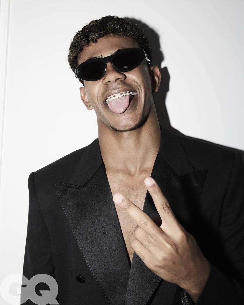 Portrait of a young adult male with curly hair wearing sunglasses and a black suit, making a peace sign against a white background.