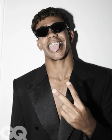 Portrait of a young adult male with curly hair wearing sunglasses and a black suit, making a peace sign against a white background.