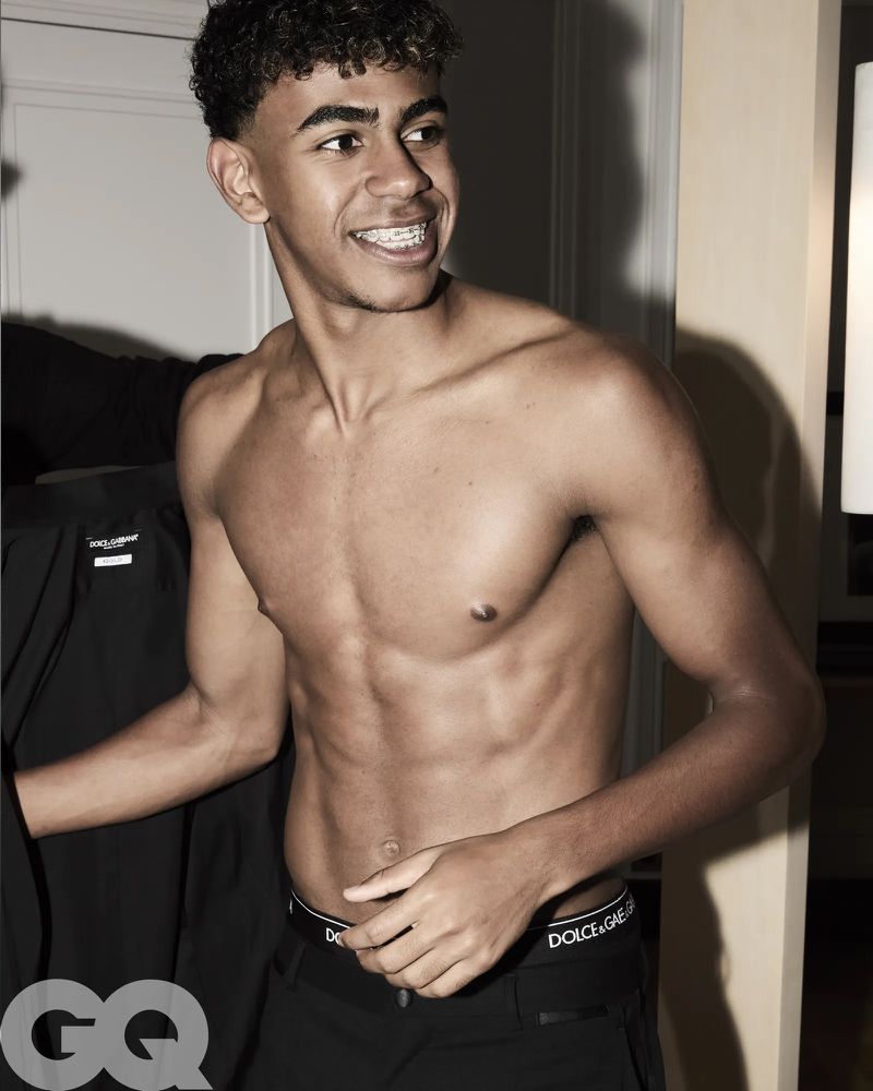 Shirtless young man with braces poses indoors against a muted wall, wearing dark pants with designer branded waistband and GQ logo on the lower left.